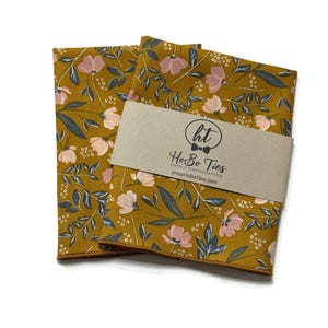 May include: Two folded fabric squares featuring a mustard yellow background with a pattern of light pink flowers, dark blue leaves, and small white "xo" text. A tan paper band wraps around the center with the text "ht HoBo Ties SIMPLY HANDCRAFTED shopHoBoTies.com".