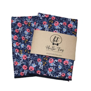 May include: Two navy blue pocket squares with a pink and white floral pattern. The pocket squares are folded and stacked on top of each other. A brown label with the text "HoBo Ties" is placed on top of the pocket squares.