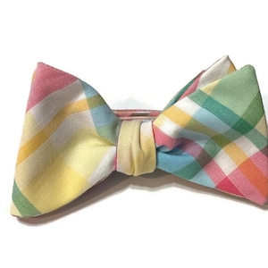May include: A colorful plaid bow tie in pastel shades of pink, yellow, green, and blue. The bow tie is made of fabric and has a pre-tied knot in the center. The bow tie is a fashion accessory.
