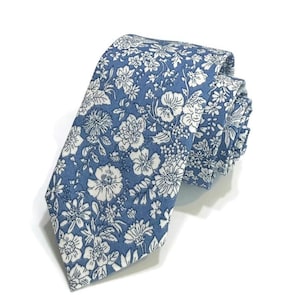 May include: A necktie featuring a blue background with a dense, white floral pattern. The fabric displays a variety of illustrated flowers and leaves in a repeating design, with the tie folded to show the pointed tip and the layered construction.