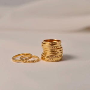 May include: A collection of gold-toned metal rings, with two lying flat and a stack of eight standing upright. Each ring features custom engraved text including names like Pomelos, Ellie, Jordan, Logan, dates in Roman numerals, and symbols.
