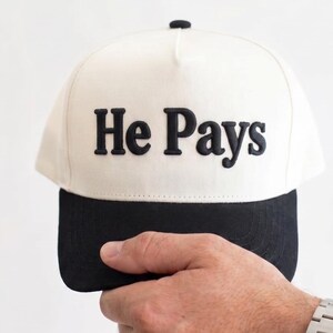 May include: A white and black baseball cap with the text "He Pays" in black block letters. The cap is held by a person wearing a silver wristwatch.