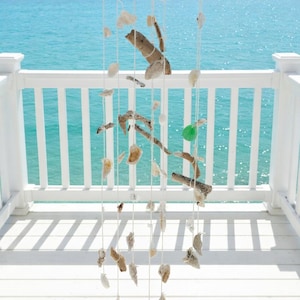 Puede incluir: Móvil decorativo de temática costera, con madera flotante, conchas marinas y vidrio de mar, suspendido de un marco rectangular de madera. Los elementos naturales están ensartados en cuerdas blancas. El fondo muestra una barandilla blanca y el océano.