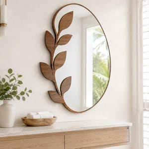 May include: An oval wall mirror featuring a dark brown wooden frame that extends into a decorative branch with several leaves along the left side. The mirror surface reflects a bright interior space with a window and green foliage visible outside.