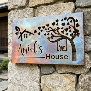 Puede incluir: Un letrero metálico rectangular con un fondo degradado azul y marrón. El letrero presenta un diseño recortado de una casa, un árbol con hojas en forma de corazón y un pájaro en una pajarera. El nombre "Amiel's House" también está recortado.