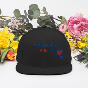 Può includere: Un cappellino snapback nero con il testo ricamato American GIRL in filo blu e rosso. A destra del testo si trova il contorno ricamato di labbra blu, un cuore rosso e un cuore blu più piccolo, il tutto centrato sul pannello frontale.