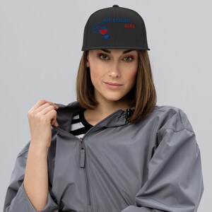 Può includere: Un cappellino da baseball nero con un disegno ricamato sul davanti. Il disegno include il testo blu "American GIRL" con un contorno blu di labbra, un cuore rosso e un piccolo cuore blu posizionati sotto il testo e le labbra.