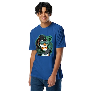 May include: A royal blue t-shirt featuring a graphic of a penguin wearing a black beanie and digital glasses, typing on a keyboard. The design includes green circuit board patterns, binary code, and a Wi-Fi symbol, all rendered in a vibrant cartoon style.