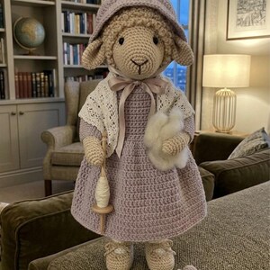May include: A crocheted sheep doll wearing a lavender dress, bonnet, and shawl, holding a spindle and a ball of yarn. The doll has beige shoes and a small crocheted spider is next to it. The doll is standing on a brown couch.