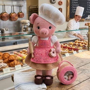 May include: A crocheted pig wearing a chef's hat and apron, holding a rolling pin and a donut. The pig has a basket of donuts and is standing in a bakery setting with pastries and a menu board.