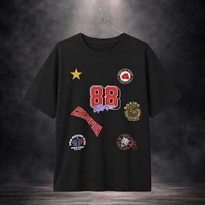 Könnte beinhalten: Schwarzes T-Shirt mit mehreren Aufnähern. Zu den Aufnähern gehören ein goldener Stern, die Zahl "88" in Rot und Blau, das Wort "Champion" in Rot und Logos zum Thema Boxen. Das T-Shirt hängt an einem Metallbügel.
