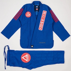 May include: A blue Gracie Barra Jiu-Jitsu uniform set. The jacket has red stripes on the shoulders and a logo on the chest. The pants also have a logo. The Adidas logo is visible on the jacket. The set is laid out on a white background.