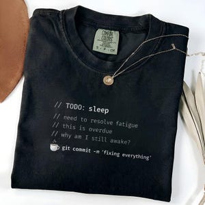 May include: Black t-shirt with white text that reads "TODO: sleep" and other related lines. A small coffee cup graphic is included. A silver necklace with a circular pendant is draped over the shirt.