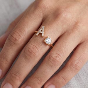 Könnte beinhalten: Ein goldfarbener Ring mit dem Buchstaben „A“, besetzt mit kleinen, klaren Steinen. Der Ring ist geteilt, mit einem runden, klaren Stein auf der anderen Seite. Der Ring befindet sich an einem Finger.