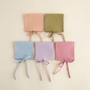 May include: Five square felt pouches in pastel colors: peach, olive green, light blue, lavender, and rose. Each pouch has a matching ribbon tie closure. The pouches are arranged in a pyramid shape against a cream background.