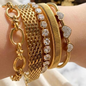 May include: A stack of five gold-toned bracelets. The set includes a chunky oval link chain, a wide woven mesh band, a single row of round clear stones in prong settings, a ribbed flat band, and a thin chain featuring three heart-shaped charms.