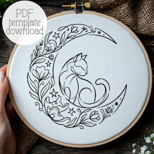 Puede incluir: Un bastidor de bordado circular de madera que contiene tela blanca con un diseño de líneas negras de un gato sentado dentro de una luna creciente rodeado de patrones florales y estrellas. El texto PDF template download es visible arriba.