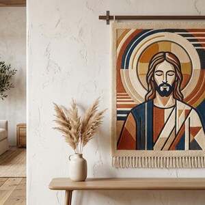 Może przedstawiać: Gobelin przedstawiający postać religijną z zamkniętymi oczami, otoczoną aureolą. Grafika zawiera geometryczne wzory w odcieniach niebieskiego, pomarańczowego i beżowego. Gobelin wisi na teksturowanej białej ścianie.
