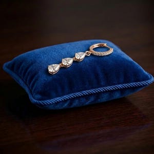 Puede incluir: Un solo pendiente de oro rosa con tres gemas claras en forma de pera y un cierre de aro. El pendiente descansa sobre un pequeño cojín cuadrado de terciopelo azul con un ribete de cuerda. El fondo es una superficie oscura de madera.