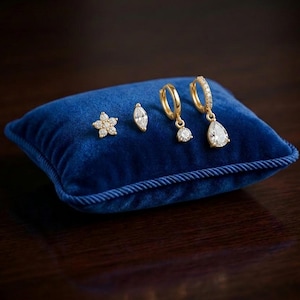 Puede incluir: Una colección de pendientes dorados expuestos sobre un pequeño cojín cuadrado de terciopelo azul. Los pendientes incluyen un pendiente de flor, un pendiente en forma de marquesa y dos pares de pendientes de aro.