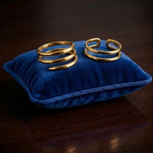 Puede incluir: Dos anillos dorados descansan sobre un pequeño cojín cuadrado de terciopelo azul. Uno tiene un diseño en espiral, el otro un diseño de doble banda. El cojín está sobre una superficie de madera oscura.
