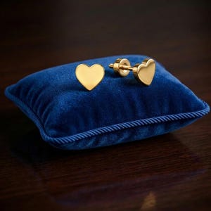 Puede incluir: Un par de pendientes de botón en forma de corazón de tono dorado con un acabado liso y pulido. Cada pendiente presenta una cara de corazón plana y un cierre de rosca diseñado para una fijación segura sobre un pequeño cojín de terciopelo azul.