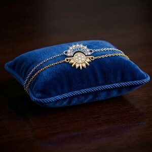 May include: A gold and silver bracelet with a sunburst design, featuring a central circle of clear stones and a half-circle of blue stones. The bracelet rests on a small, square, blue velvet pillow with a twisted rope border.
