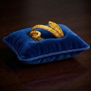 Può includere: Un braccialetto color oro a forma di serpente poggia su un piccolo cuscino quadrato di velluto blu con bordatura a corda. Il braccialetto presenta un motivo strutturato a scaglie e un design a spirale. Il cuscino è appoggiato su una superficie di legno scuro.