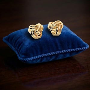 Pode incluir: Um par de brincos de tarracha em tom dourado polido com um design de nó de amor tridimensional. Cada brinco é feito com laços entrelaçados suaves que criam uma forma arredondada e escultural com um acabamento metálico de alto brilho.