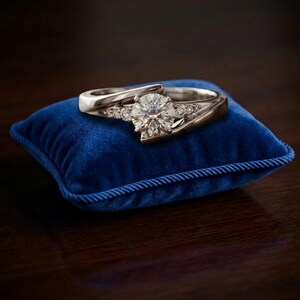 Puede incluir: Un anillo de compromiso plateado con una gema grande, redonda y transparente y piedras de acento más pequeñas. El anillo está colocado sobre un pequeño cojín cuadrado de terciopelo azul con un ribete de cordón decorativo.