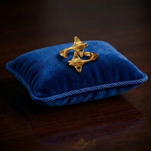 Puede incluir: Un anillo de tono dorado que presenta dos formas estilizadas y puntiagudas similares a alas que se encuentran en el centro. El anillo se muestra sobre un pequeño cojín cuadrado de terciopelo azul intenso con bordes ribeteados y acabado metálico.
