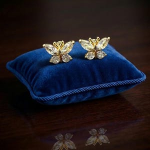 Puede incluir: Un par de pendientes de mariposa dorados con detalles claros y brillantes. Los pendientes se exhiben sobre un pequeño cojín cuadrado de terciopelo azul con ribete de cuerda. El fondo es una superficie oscura y reflectante.