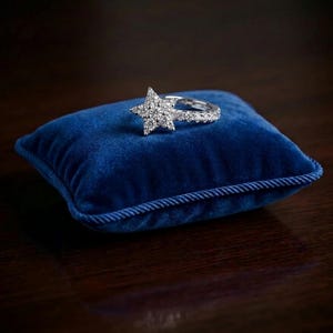 Puede incluir: Un anillo de diamantes con forma de estrella descansa sobre un pequeño cojín cuadrado de terciopelo azul con ribete de cuerda. El anillo presenta un grupo de diamantes en forma de estrella y una banda adornada con diamantes más pequeños.