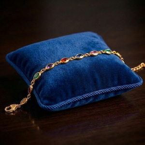 Puede incluir: Una pulsera dorada con gemas ovaladas de colores descansa sobre un cojín de terciopelo azul. La pulsera presenta una serie de gemas vibrantes en rojo, verde, morado y azul, unidas por una delicada cadena dorada. El cojín es cuadrado y tiene un ribete de cuerda.