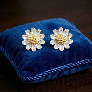 Puede incluir: Un par de pendientes de botón con forma de margarita, con pétalos plateados que irradian desde un centro dorado texturizado. Los pendientes se muestran uno al lado del otro, exhibiendo un diseño floral detallado con un acabado metálico.