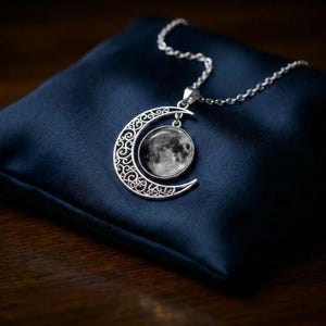 Puede incluir: Un collar de tono plateado con un colgante de media luna con intrincados recortes de filigrana. Un inserto circular de cabujón muestra una imagen fotográfica detallada en blanco y negro de la superficie lunar, suspendido de una cadena plateada.