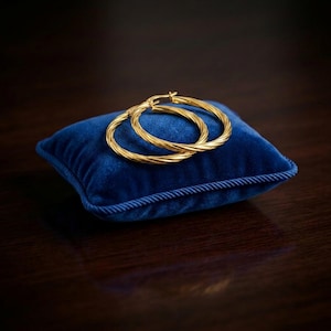 Puede incluir: Un par de pendientes de aro dorados con una textura de cuerda trenzada. Los pendientes tienen forma circular con un acabado pulido y descansan sobre un pequeño cojín cuadrado de terciopelo azul profundo con borde trenzado.