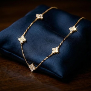 Puede incluir: Un collar dorado con cinco colgantes de trébol de cuatro hojas, cada uno con pequeñas gemas brillantes. El collar descansa sobre una superficie de satén azul oscuro, creando un contraste que resalta los detalles de la joya.