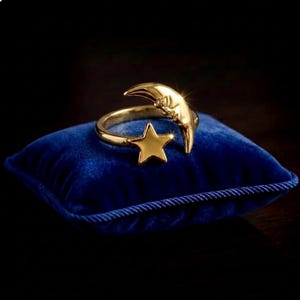 Puede incluir: Un anillo abierto de tono dorado que presenta una luna creciente con un rostro detallado en un extremo y una estrella de cinco puntas en el otro. El anillo tiene un acabado metálico pulido y descansa sobre un cojín cuadrado de terciopelo azul.