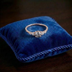 Può includere: Un anello in oro rosa con una pietra centrale a taglio rotondo affiancata da due pietre rotonde più piccole su ciascun lato. L'anello è posizionato su un morbido cuscino in velluto blu intenso con bordo intrecciato, mostrando una fascia lucida.