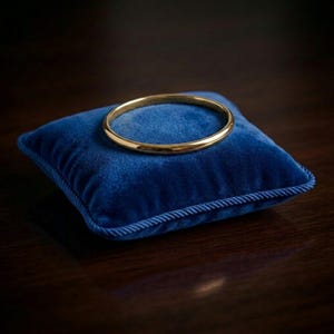 Puede incluir: Una pulsera dorada descansa sobre un pequeño cojín cuadrado de terciopelo azul con ribete de cuerda. La pulsera es circular y parece ser de metal. El cojín es de color azul vibrante, y el fondo es una superficie de madera oscura.