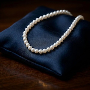 Puede incluir: Un delicado collar de perlas descansa sobre un cojín de satén azul oscuro. El collar está compuesto por numerosas perlas blancas, pequeñas y redondas. El cojín de satén tiene una superficie lisa y reflectante.