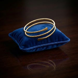 Puede incluir: Un brazalete dorado con textura martillada y diseño en espiral descansa sobre un pequeño cojín cuadrado de terciopelo azul. El brazalete tiene un diseño abierto único y se exhibe sobre una superficie de madera oscura.