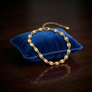 Puede incluir: Una pulsera dorada con eslabones ovalados, cada uno con una pequeña perla blanca redonda. La pulsera se exhibe sobre un pequeño cojín cuadrado de terciopelo azul. El fondo es una superficie oscura y reflectante.