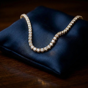 Puede incluir: Un collar de tenis dorado que presenta una fila continua de piedras redondas, transparentes y facetadas engastadas en un marco metálico. El collar está dispuesto en forma curva, mostrando el tamaño y la disposición uniforme de las piedras.