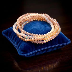 Puede incluir: Una pila de tres pulseras circulares con pequeñas cuentas redondas en tonos melocotón, crema y vidrio translúcido iridiscente. Las cuentas están ensartadas en un bucle continuo, creando una apariencia en capas con texturas variadas.
