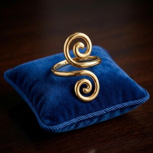 Puede incluir: Un anillo de tono dorado que presenta un diseño de doble espiral con dos extremos redondeados y enrollados que se extienden desde una banda central. El metal tiene un acabado liso y pulido que refleja la luz, creando un aspecto metálico brillante.