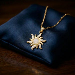 Puede incluir: Collar dorado con colgante de sol. El colgante tiene una piedra blanca central, rodeada de cristales transparentes y rayos de sol. Se muestra sobre un cojín de satén azul oscuro.
