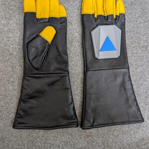 May include: A pair of black leather gloves with yellow fingertips and a gray and blue emblem on the back of one glove. The gloves have long cuffs and are likely for protection or a costume.