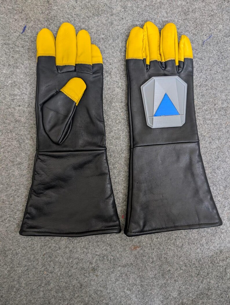 May include: A pair of black leather gloves with yellow fingertips and a gray and blue emblem on the back of one glove. The gloves have long cuffs and are likely for protection or a costume.